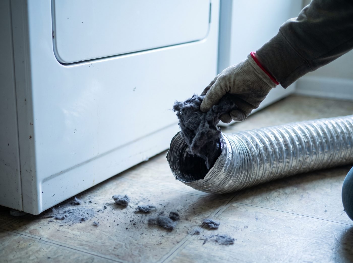 A gloved hand pulling a thick wad of lint from a disconnected dryer vent duct