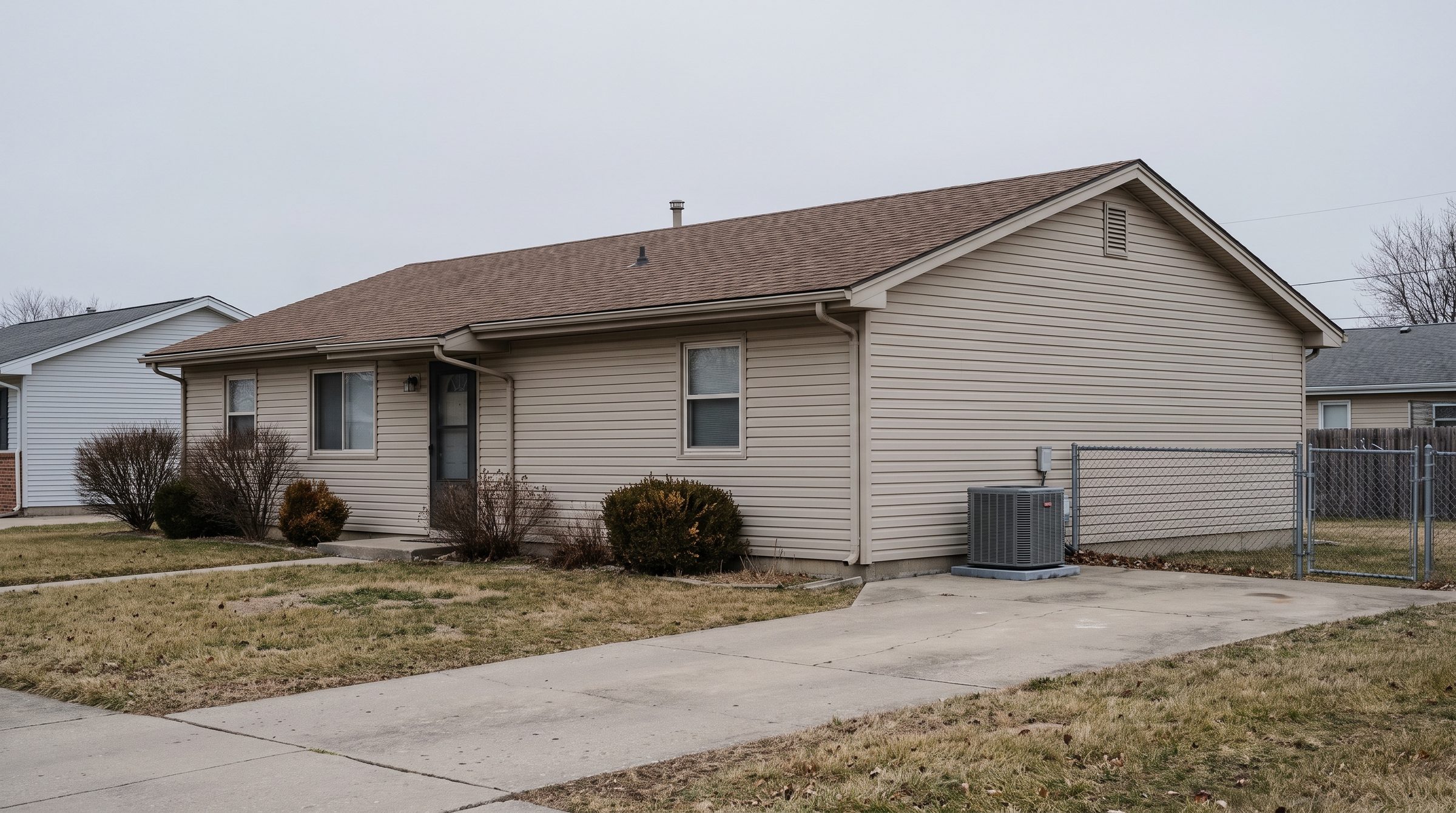 The front of an ordinary single-storey suburban house on an overcast day, condenser unit at the side